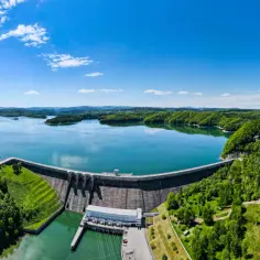 Zapora wodna z elektrownią, otoczona zielonymi wzgórzami i błękitnym jeziorem.