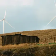 Dwie białe turbiny wiatrowe na wzgórzu nad drewnianą szopą.