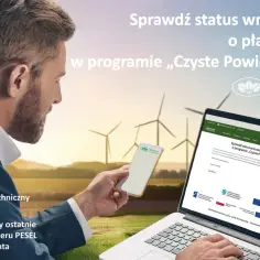 Mężczyzna sprawdza status wniosku na laptopie i telefonie, na tle farmy wiatrowej.