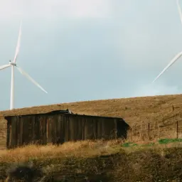Dwie białe turbiny wiatrowe górują nad suchym, trawiastym wzgórzem, gdzie stoi stary, drewniany budynek. To nowoczesna elektrownia wiatrowo słoneczna.